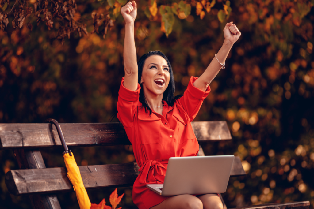 Woman celebrates success with laptop outdoors in autumn.