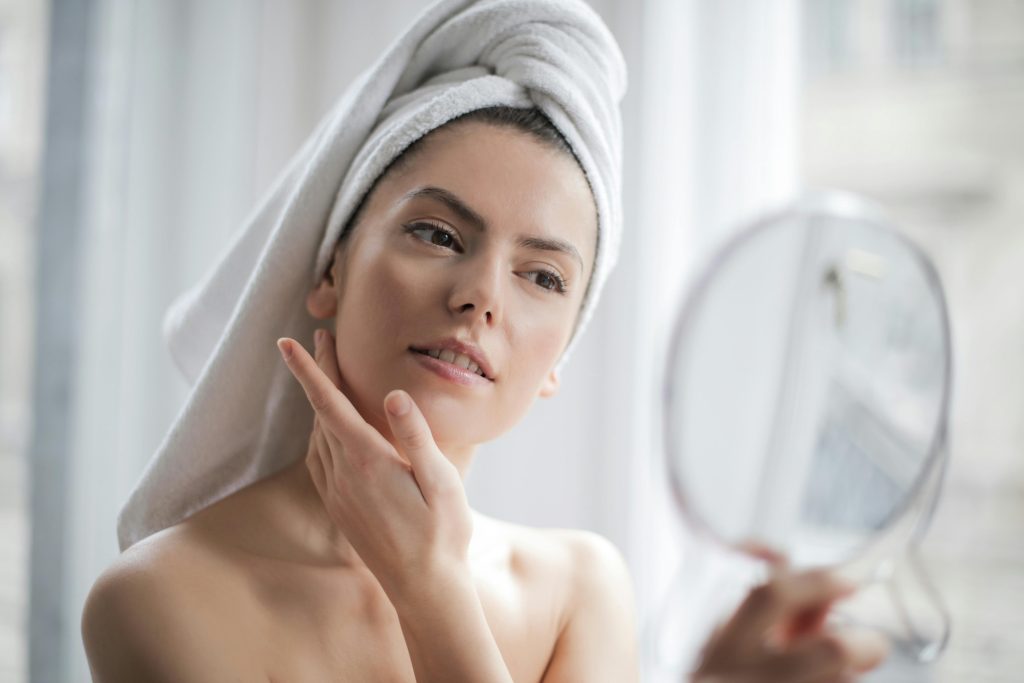 Woman with towel examines skin in mirror.